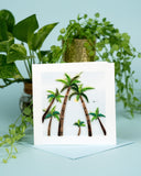 Quilled Palm Trees Greeting Card standing on top of blue envelope on blue table in front of green plants
