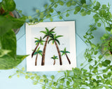 Quilled Palm Trees Greeting Card on blue background with blue envelope laying on table surrounded by green plants