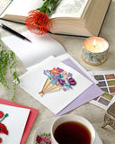 Quilled Playful Flower Bouquet Greeting Card laying on a knitted cloth next to a book, flowers, candles, a teacup, and stamps.