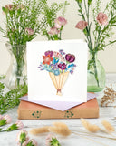 Quilled Playful Flower Bouquet Greeting Card sitting on a book on top of a wooden table, next to pink flower vases and dry flowers.