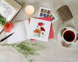 Quilled Red & Orange Poppies Greeting Card laying on linen cloth, next to stamps, teacups, flowers, a candle, and an open book.