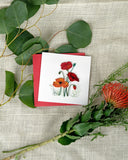 Quilled Red & Orange Poppies Greeting Card laying on linen cloth next to leaves and an orange flower.
