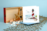 Quilled Red & White Lighthouse Greeting Card standing up on top of a rope net and in front of a wooden box with seashells in front of a light blue background.