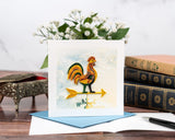 quilled rooster weathervane greeting card with blue envelope next to card insert with pen, flowers, and books in front of white background