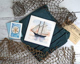 Quilled Schooner at Sunset Greeting Card laying on a blue napkin, besides a Quilled Sand Dollar Gift Enclosure Mini Card, surrounded by net.