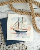 Quilled Schooner at Sunset Greeting Card laying on a white wooden surface, resting on a net, next to a rope.