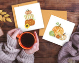Quilled Stacked Pumpkins Greeting Card on wooden table next to quilled pumpkin card with sweater, leaf, hand holding coffee