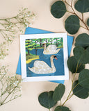 Quilled Two Swans Greeting Card with blue envelope on beige background next to eucalyptus and baby's breath florals