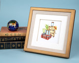 quilled vintage luggage greeting card inside of gold frame next to books in front of light blue background