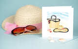 Quilled Vitamin Sea Greeting Card standing up in front of a woven beach hat and a light blue background while covered in seashells.