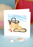 Quilled Vitamin Sea Greeting Card standing up in front of a woven beach hat, a pink Hand-woven Bamboo Bag, and a light blue background while covered in seashells.