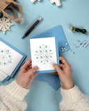 Quilled Snowflake Note Card Box Set card 1 being held over over a light blue background, blue envelopes, wax melts, and a Quilled Snowflake Ornament.