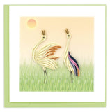 Two white cranes standing in a green field in front of an orange sky.