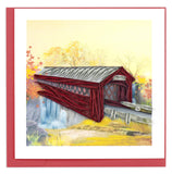 A blank greeting card of a quilled red covered bridge over a watercolor scene.